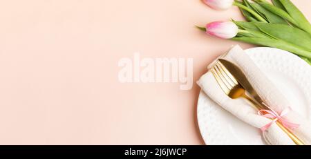 Elegance romantic table setting with golden cutlery on white plate and pink tulips on pastel pink backdrop. Festive banner. Holiday background. Top vi Stock Photo