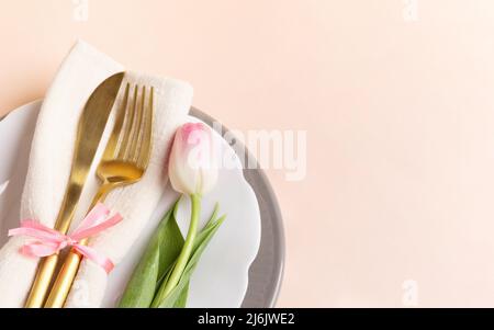 Elegance romantic table setting with golden cutlery on plates and one pink tulip on pastel pink backdrop. Festive banner. Holiday background. Top view Stock Photo