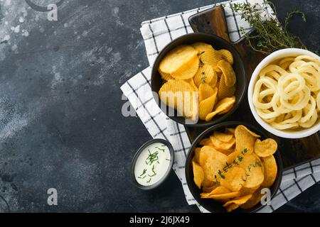 Unhealthy food. Snacks. All classic potato snacks with peanuts, popcorn and onion rings and salted pretzels in bowl plates on black concrete backgroun Stock Photo