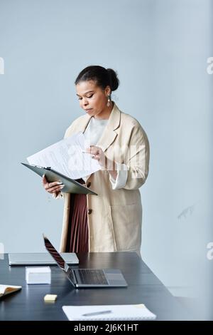 Vertical portrait of elegant black businesswoman reading document while standing in office against minimal grey background Stock Photo