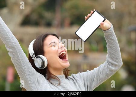 Happy teen singing showing smart phone blank screen in a park Stock Photo