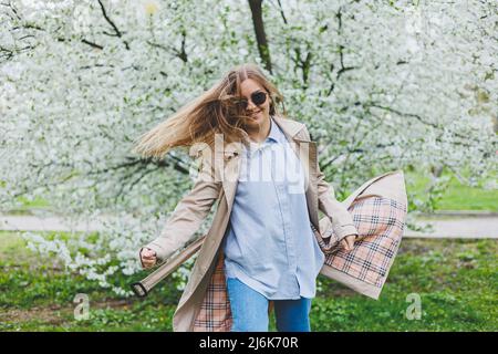 A young woman with blond hair enjoys a blooming spring garden. Travel, spring break. Fashionable style. A woman in sunglasses and a beige trench coat Stock Photo