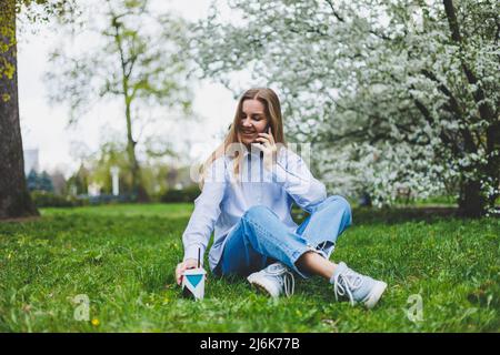 Top view of woman sitting in park garden on the green grass with smartphone, headphones, tablet, book and coffee in the hand Stock Photo