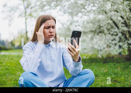 Young emotional asian woman using mobile phone watching video online, rearing news sitting on grass. Excited female shopping online Stock Photo