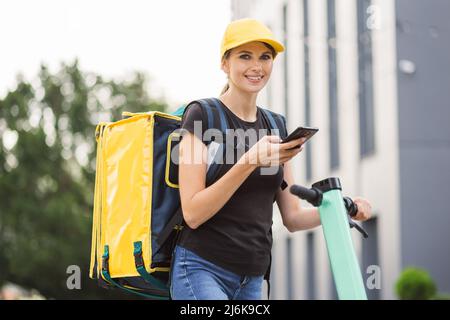 Side view of delivery woman taking order on cellphone standing near electric scooter on urban playground. Female courier using mobile app during work with customers in summer city stree Stock Photo