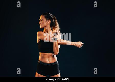 Young muscular woman stretching her arm in studio on black background looking away Stock Photo