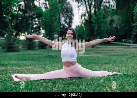 Attractive young brunette in nature. Beautiful Caucasian woman does yoga in splits pose with her hands raised to side in park on summer day Stock Photo