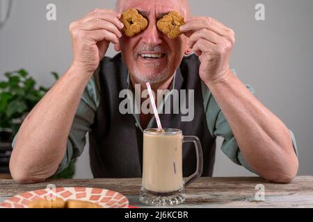 casual dressed mature man with two cookies on his eyes sitting at a table drinking iced coffee through a straw Stock Photo