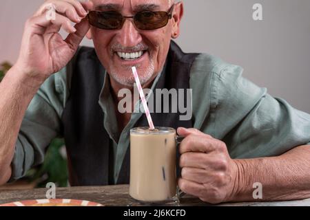 casual dressed mature man with sunglasses sitting at a table drinking iced coffee through a straw Stock Photo