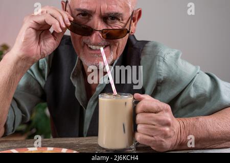 casual dressed mature man with sunglasses sitting at a table drinking iced coffee through a straw Stock Photo