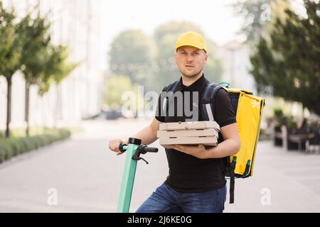 Attractive delivery man holding stack of pizza cardboard boxes using electric scooter waiting for a client on the background of modern buildings. Male courier with pizza looking at camera Stock Photo