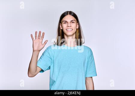 Positive teenage guy waving hand, looking at camera, light studio background Stock Photo
