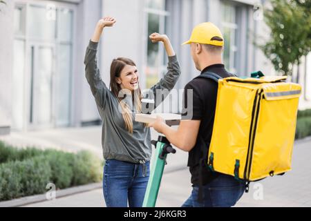 Delivery man giving pizza to happy customer. Food delivery courier giving order while standing on electric scooter wearing company yellow backpack and cap. Delivery man giving lunch to client. Stock Photo