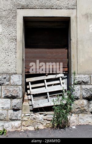 Old dilapidated wooden boards entrance doors partially covered with broken rusted metal blinds locked with rusted padlock on top of stacked Stock Photo