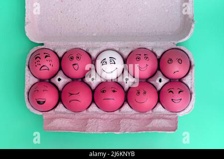 Painted red eggs in a cardboard tray top view. Eggs painted with emotional faces and a white egg. Ten eggs scattered on a green background top view. W Stock Photo