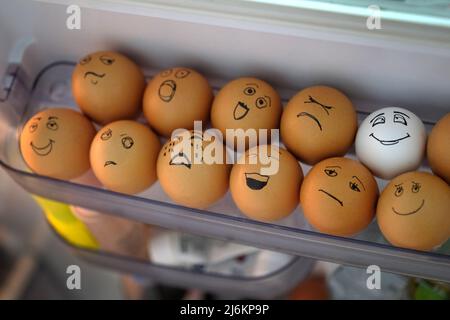 Brown eggs and one white in a plastic container in the refrigerator. Eggs in the refrigerator with faces of different emotions. The concept of a man i Stock Photo