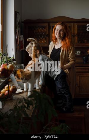 Ginger haired woman on kitchen with child and cat looking in laptop. Vintage house, cozy family life. Boy with mother at home.  Stock Photo