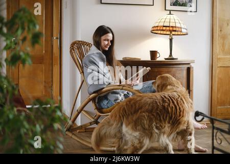 Young woman reading book with dog Golden retriever, sitting on rocking chair. Cozy home atmosphere, slow living, people with pets. Vintage house Stock Photo