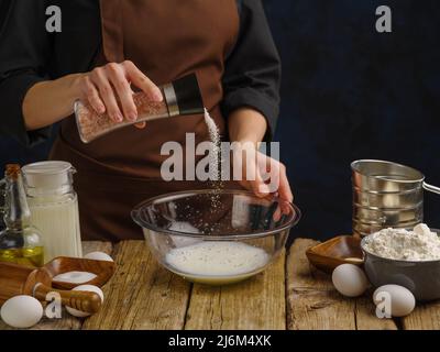 A chef in a dark uniform prepares dough in a glass bowl on a wooden kitchen table. Sprinkle salt into the dough. Salt in frozen flight. There are a lo Stock Photo