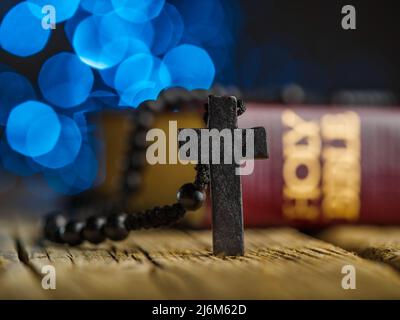 Rosary with a crucifix and the Holy Bible on a wooden table, dark blue background with lights. Symbols of Christianity, Catholicism. Religion, faith, Stock Photo