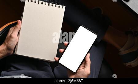 Close-up image, Male hands holding empty spiral notepad and cellphone white screen mockup, top view Stock Photo