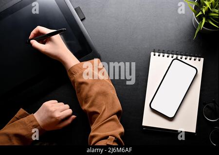 Millennial female graphic designer using graphic tablet with stylus pen, designing her graphic work at office desk with accessories and smartphone moc Stock Photo