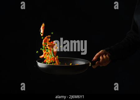 A professional chef prepares shrimps with vegetables in a frying pan on a black background. Frozen in-flight food. Seafood cooking. Healthy vegetarian Stock Photo