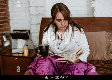 Hispanic senior mature woman reading a book in bed at home in Mexico Latin America Stock Photo