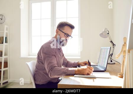 Businessman in glasses sitting at desk, working on laptop computer and taking notes Stock Photo
