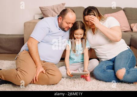 Happy family of three persons sitting on the carpet at home with and their girl holding a paper in her hand together looking at it passing weekend Stock Photo