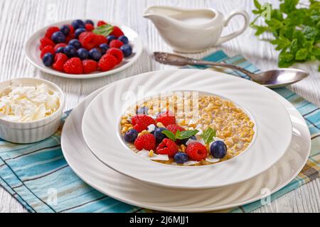 hulled oat porridge cooked with coconut milk topped with fresh raspberries, blueberries and coconut chips in white bowl on wood table, horizontal view Stock Photo
