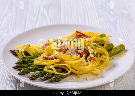 roasted asparagus spears with pasta sprinkled with shredded pecorino cheese, fried bacon pieces and parsley on white plate on white textured wood tabl Stock Photo