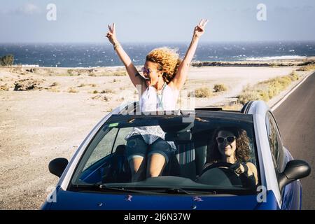 Two young women traveling in a convertible car and having fun during journey. Young woman posing with v sign while friend driving car on road. Two