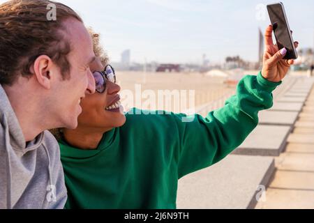 young latin woman smiling with her friend while taking a selfie photo with the mobile phone, concept of lifestyle and friendship Stock Photo