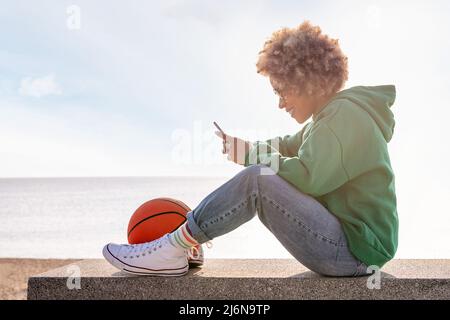 smiling young latin woman in a hoodie sitting on a bench by the beach with a basketball using her phone, concept of lifestyle and technology of commun Stock Photo