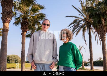 young caucasian man and latin woman smiling happy looking at camera next to palm trees, concept of friendship and couple relationship Stock Photo