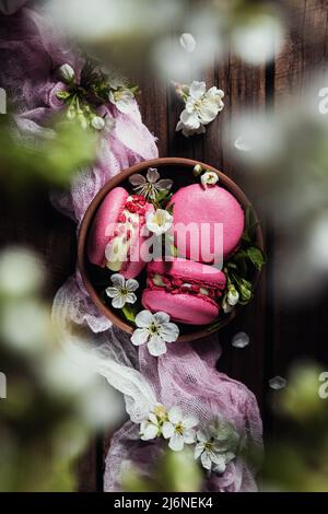 Top view on pink strawberry macaron cookies in bowl on wooden brown deck boards background, white apple blossom flowers. Beautiful colorful composition flat lay with sweet small French macaroon cakes Stock Photo