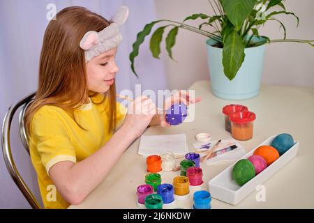 A child paints Easter eggs with paints. Portrait of caucasian girl, child 9-10 years old decorating easter eggs in the domestic kitchen. The concept o Stock Photo
