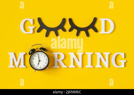 Creative good morning concept. White letters, black eyeleshes and alarm clock on yellow background. Top view, Flat lay. Greeting card. Stock Photo