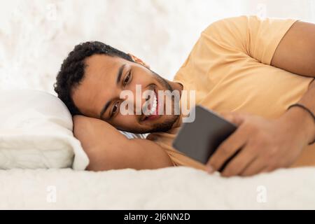 Happy Arabic Guy Texting Via Phone Lying In Bedroom Stock Photo