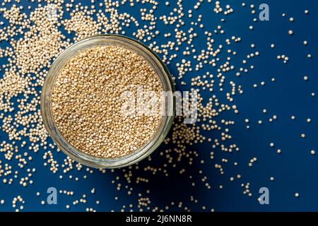 Raw quinoa in a glass jar and scattered grains over blue background. Superfood chenopodium quinoa for gluten free dieting. White quinoa seeds ingredie Stock Photo