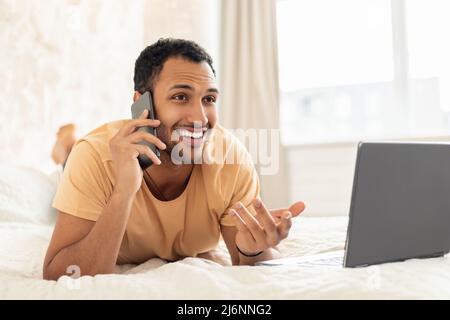 Middle Eastern Guy Talking On Cellphone Using Laptop In Bedroom Stock Photo