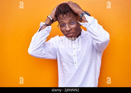 Shaken African American male in shirt and glasses touching head and looking at camera against orange background Stock Photo