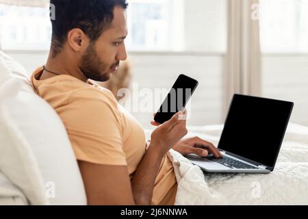 Man Using Phone And Laptop Sitting In Bed At Home Stock Photo