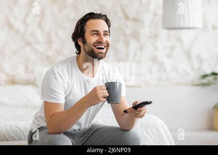 Joyful guy sitting on bed, watching TV and drinking coffee Stock Photo