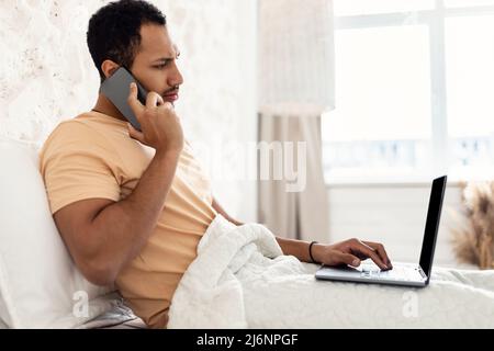 Arabic Guy Talking On Phone Using Laptop Working In Bedroom Stock Photo