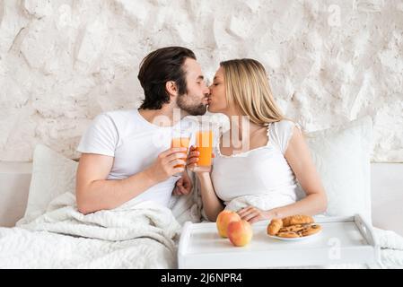 Loving young couple having breakfast in bed together, kissing, spending honeymoon at hotel Stock Photo