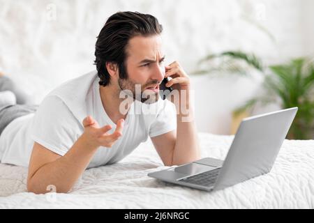 Furious man making phone call and using laptop in bed Stock Photo
