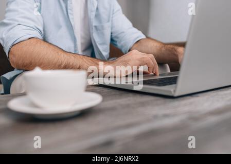 Hands of modern programmer or office manager over laptop keyboard under work on business plan. A guy in a blue shirt and white T-shirt. Cup of coffee on the table. Freelance Stock Photo