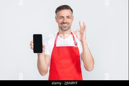 happy mature man in apron presenting smartphone with copy space. ok Stock Photo